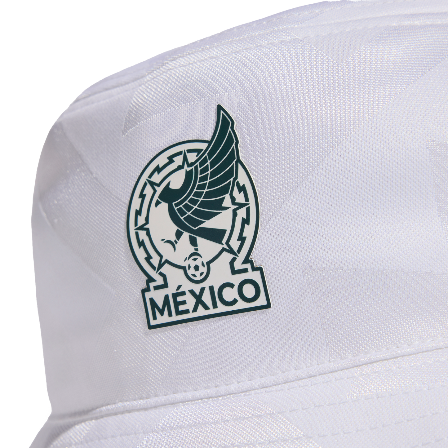 A close-up of a white bucket hat featuring a dark green and white patch. The patch depicts a stylized Aztec eagle, a soccer ball, and the word "MÉXICO" at the bottom.