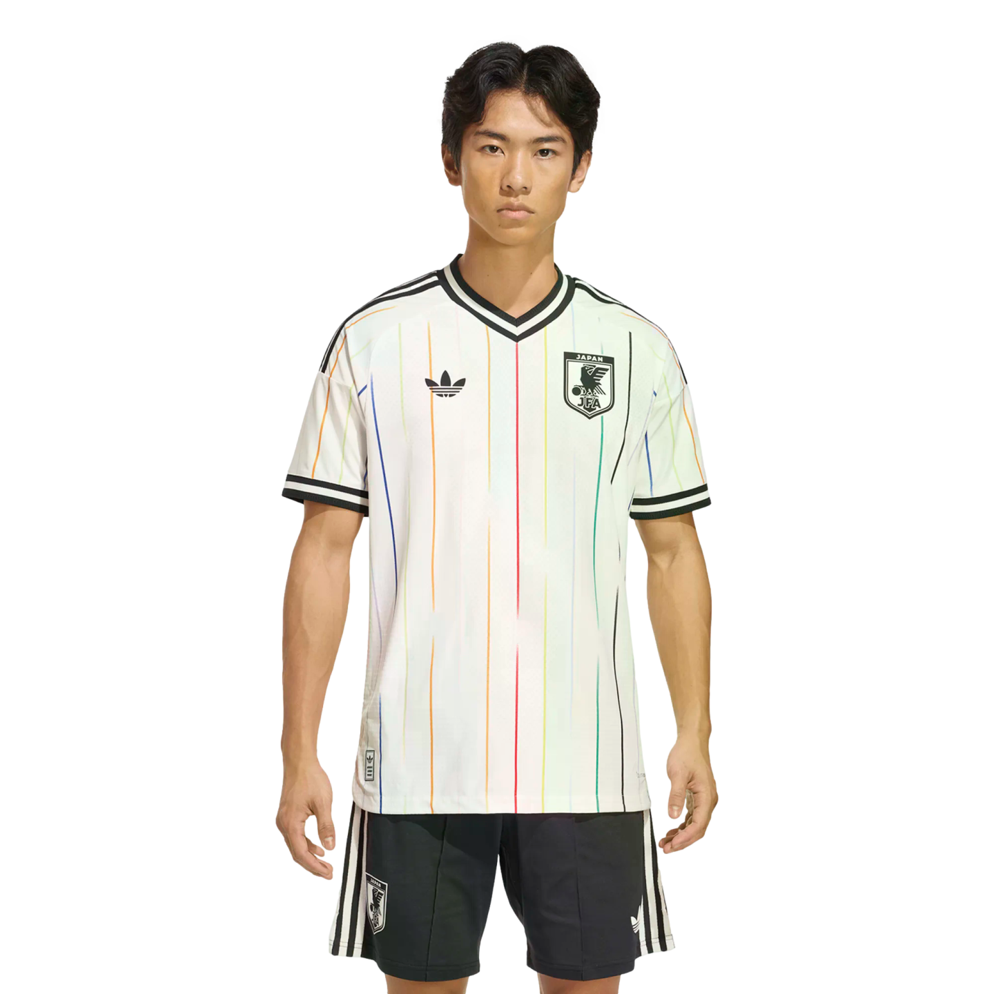 A man models a white Japan football jersey featuring thin vertical rainbow stripes and matching black shorts against a solid black background.