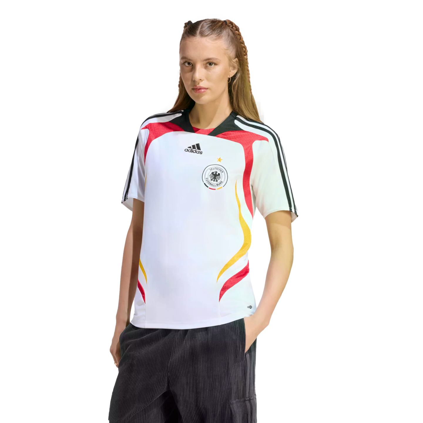 A woman in a white Germany national team soccer jersey and dark pants poses against a white background. Her hair is long and braided. Her hands are in her pockets as she looks at the camera.