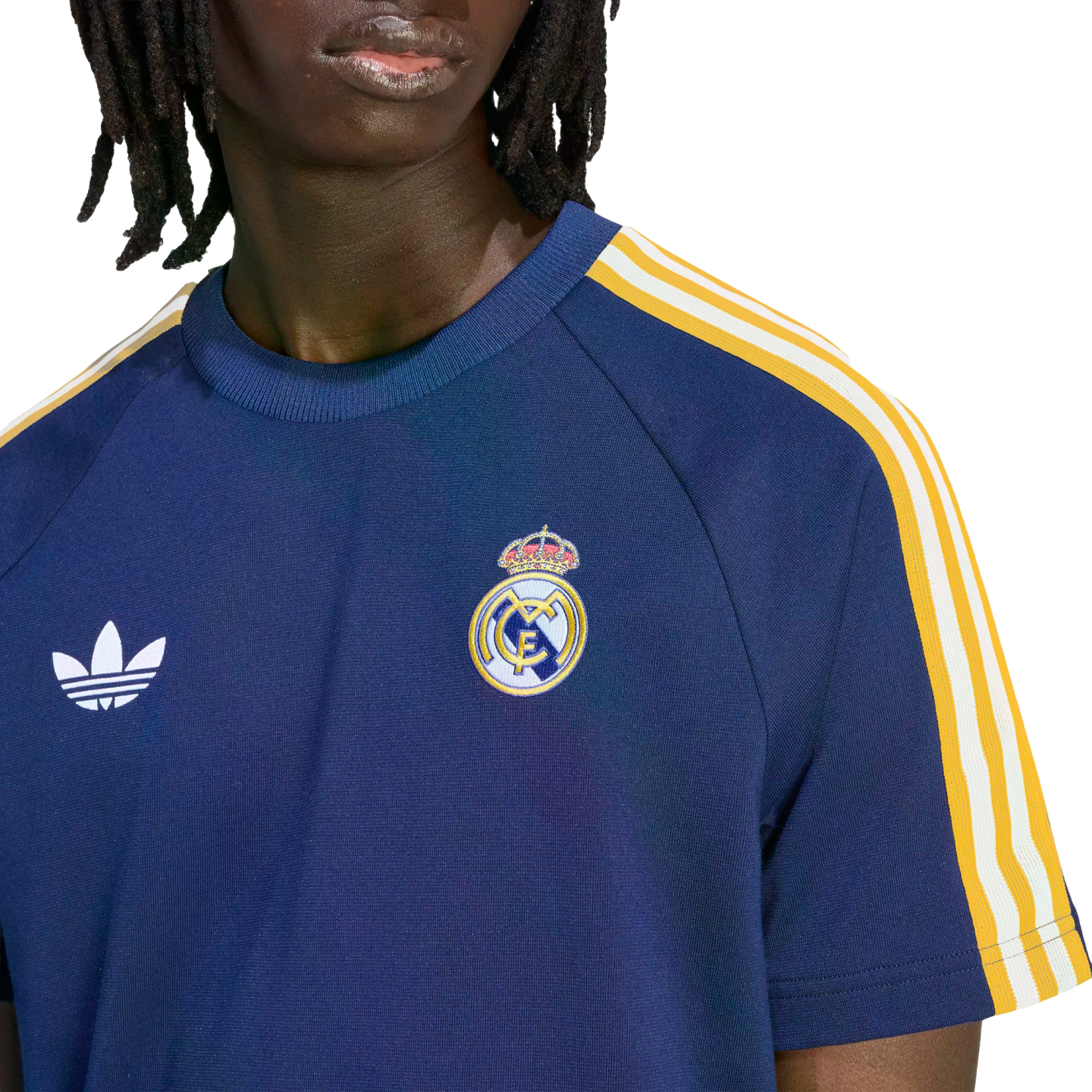 A person with dreadlocks wears a navy blue Real Madrid shirt featuring a white Adidas logo and the club's crest. The sleeves have yellow and white stripes.