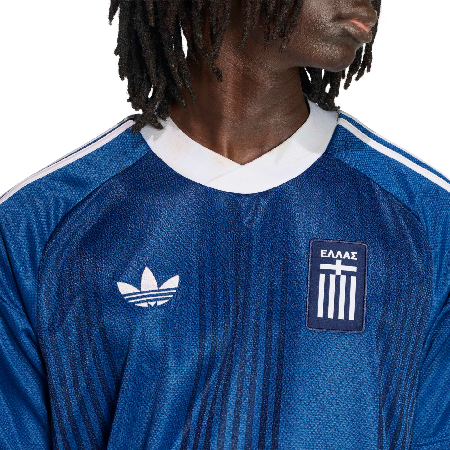 Close-up of a person wearing a blue Adidas Greece soccer jersey with a white trefoil logo and "ΕΛΛΑΣ" crest against a black background.