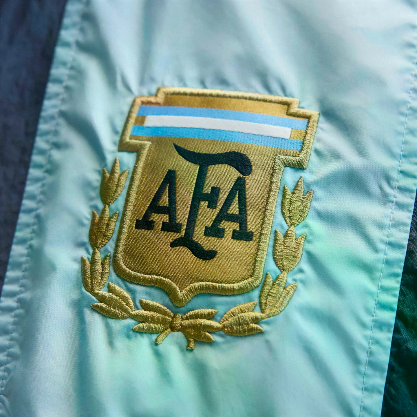 Close-up of the embroidered gold Argentine Football Association (AFA) shield logo on light blue fabric, featuring "AFA" in dark letters and a laurel wreath.