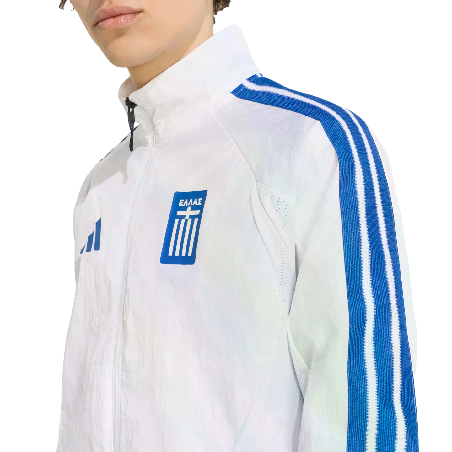 A person wearing a white track jacket with blue sleeve stripes and a blue Greek national team 'ΕΛΛΑΣ' crest on the chest.