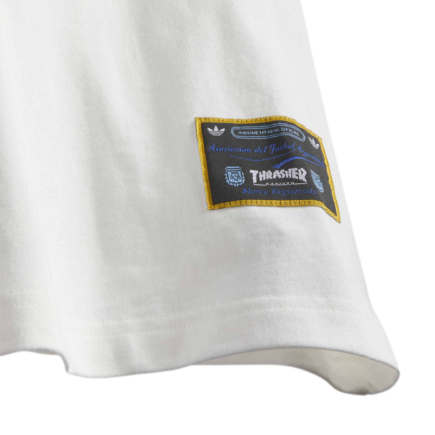 Close-up of a white shirt hem featuring a black and gold woven patch with logos for Adidas, Thrasher magazine, and the Argentine Football Association (AFA).