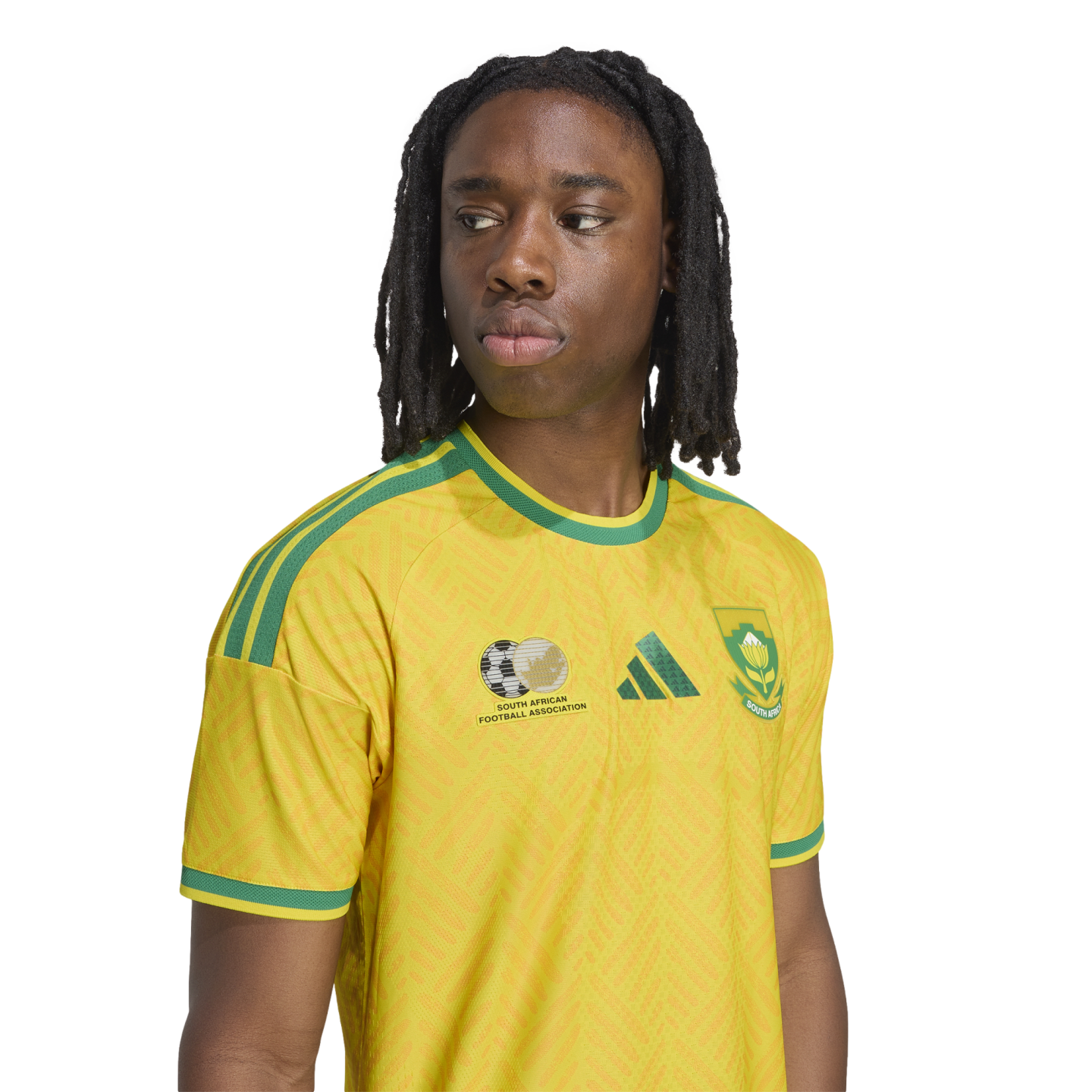 A person with long dreadlocks wearing a yellow and green South African national soccer jersey, looking to the side against a white background.