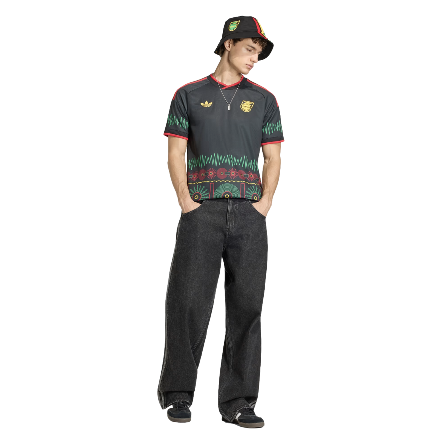 A man in a black Adidas Jamaica jersey with colorful patterns, matching bucket hat, and wide-leg dark jeans stands against a black background.