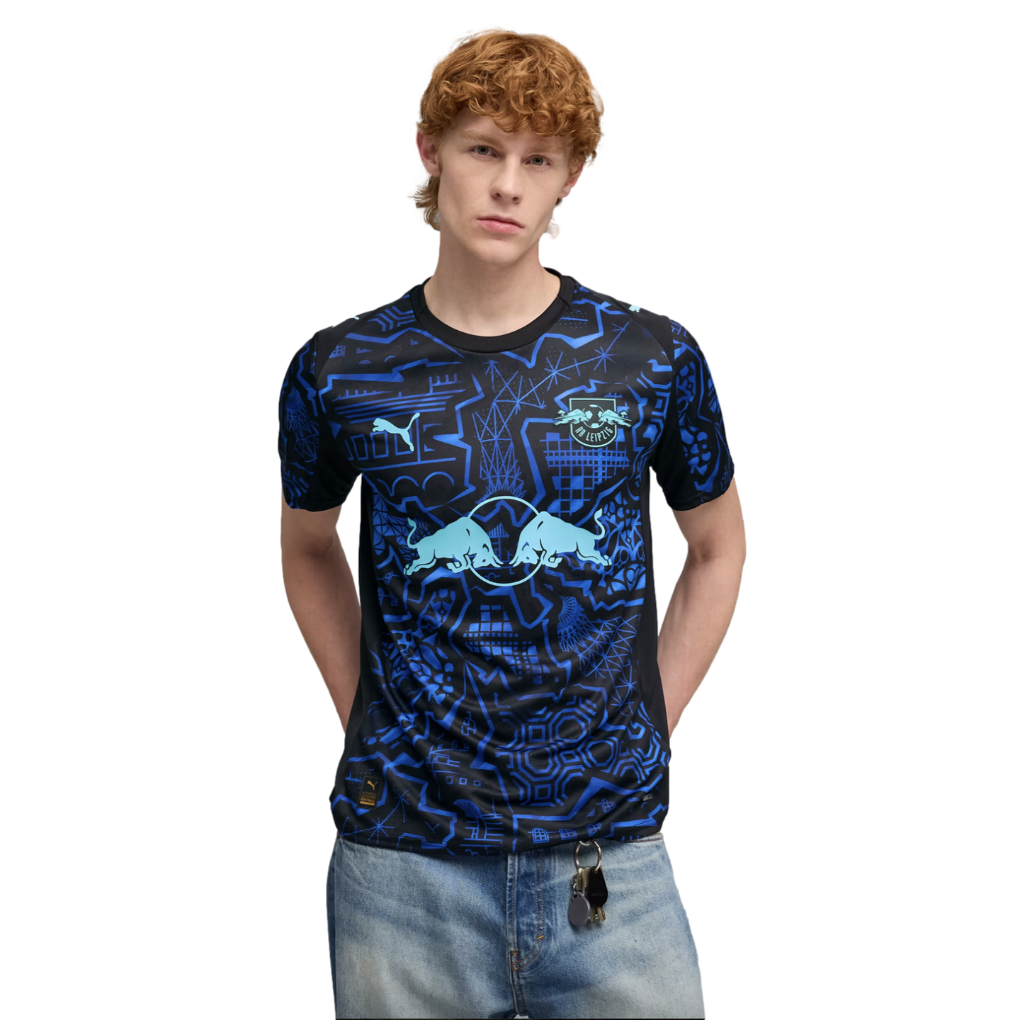 A man with red hair wearing a black and blue patterned Puma RB Leipzig soccer jersey and light blue jeans.
