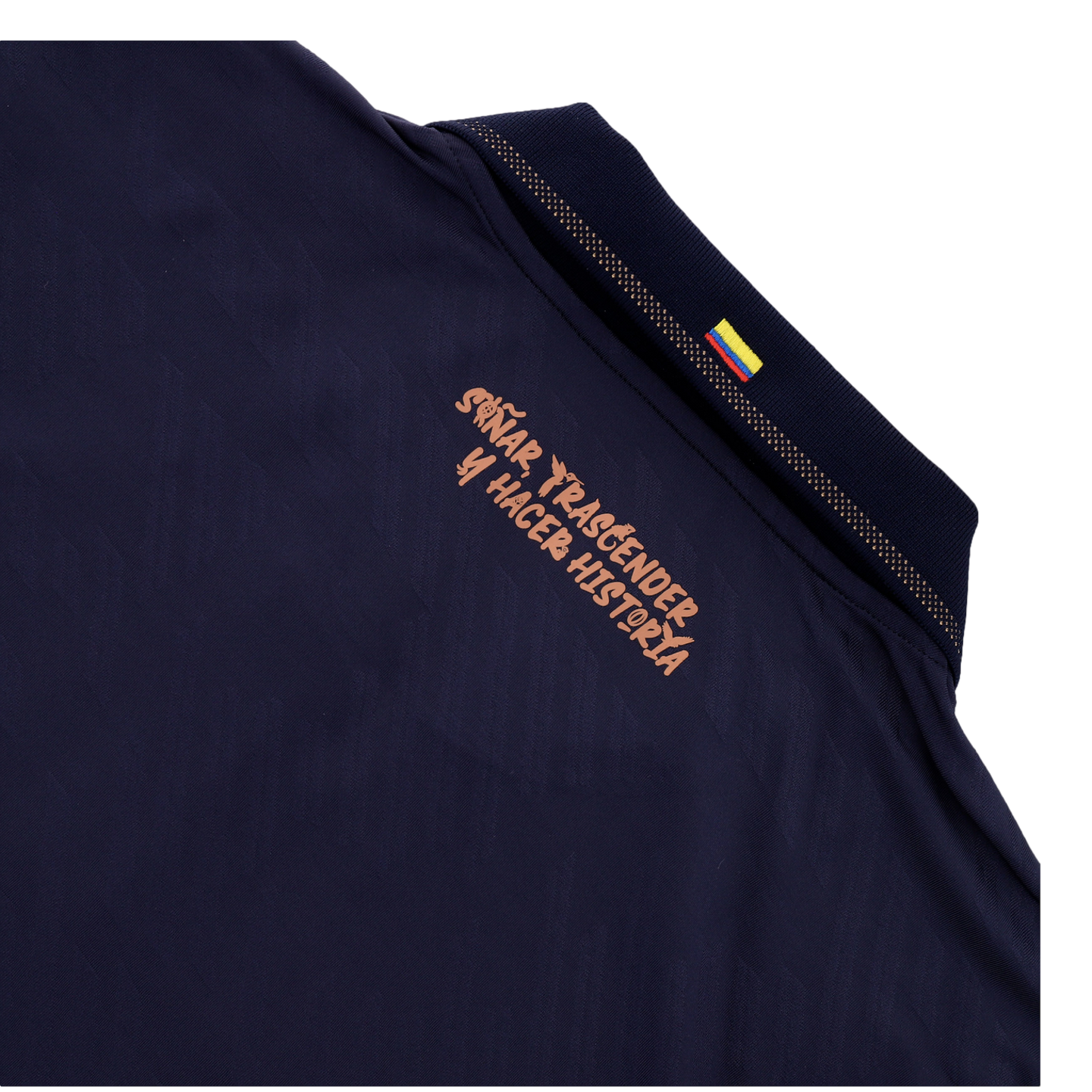 A close-up of the back collar area of a dark navy blue shirt. Below the collar, a Spanish slogan is printed in a stylized copper-colored font that reads "SOÑAR, TRASCENDER Y HACER HISTORIA." A small Colombian flag is visible on the ribbed collar, and the 