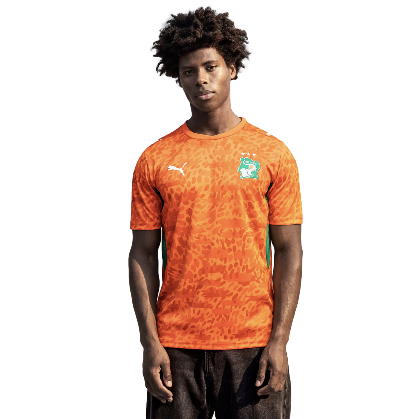 A medium shot of a young man with dark, curly hair wearing an orange Puma soccer jersey for the Côte d'Ivoire national team. The jersey features an all-over tonal leopard-print pattern, a green and white national crest on the left chest, and a white Puma 
