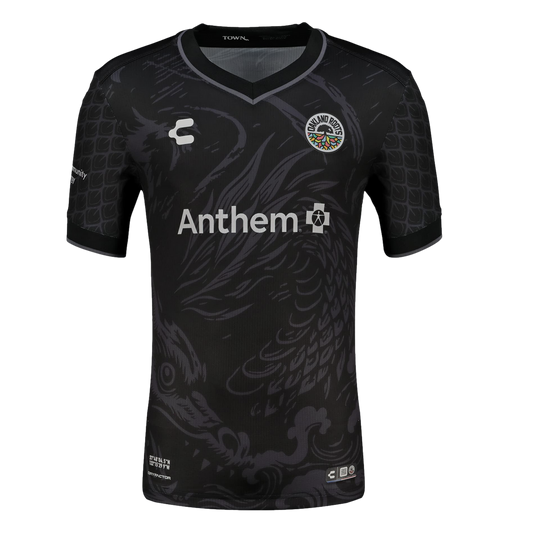 A black short-sleeved soccer jersey featuring a large, subtle grey dragon illustration across the front. The jersey includes the colorful Oakland Roots SC crest on the chest, a white Anthem sponsor logo in the center, and a repeating grey scale pattern on