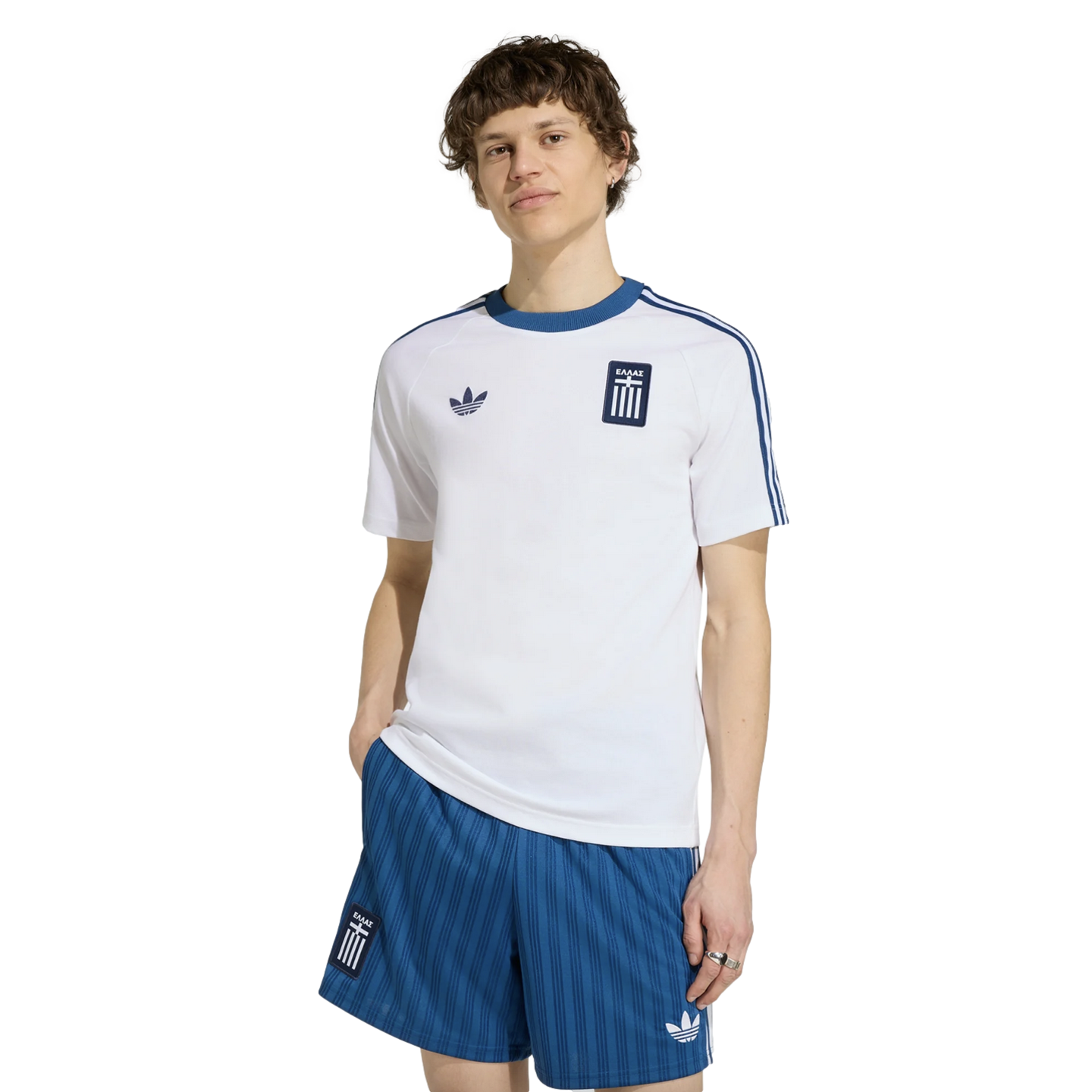 A man models a white Adidas t-shirt and blue striped shorts, both with the Greek national team crest, against a black background.