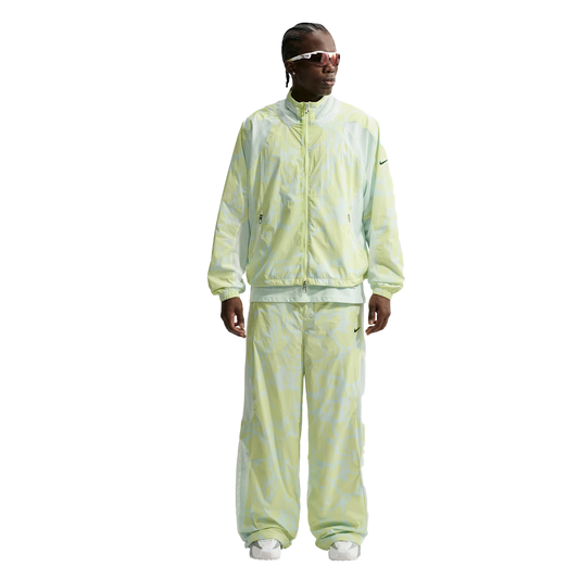 A person wearing a pale green patterned Nike tracksuit with wide-leg pants, white sunglasses, and white sneakers against a white background.