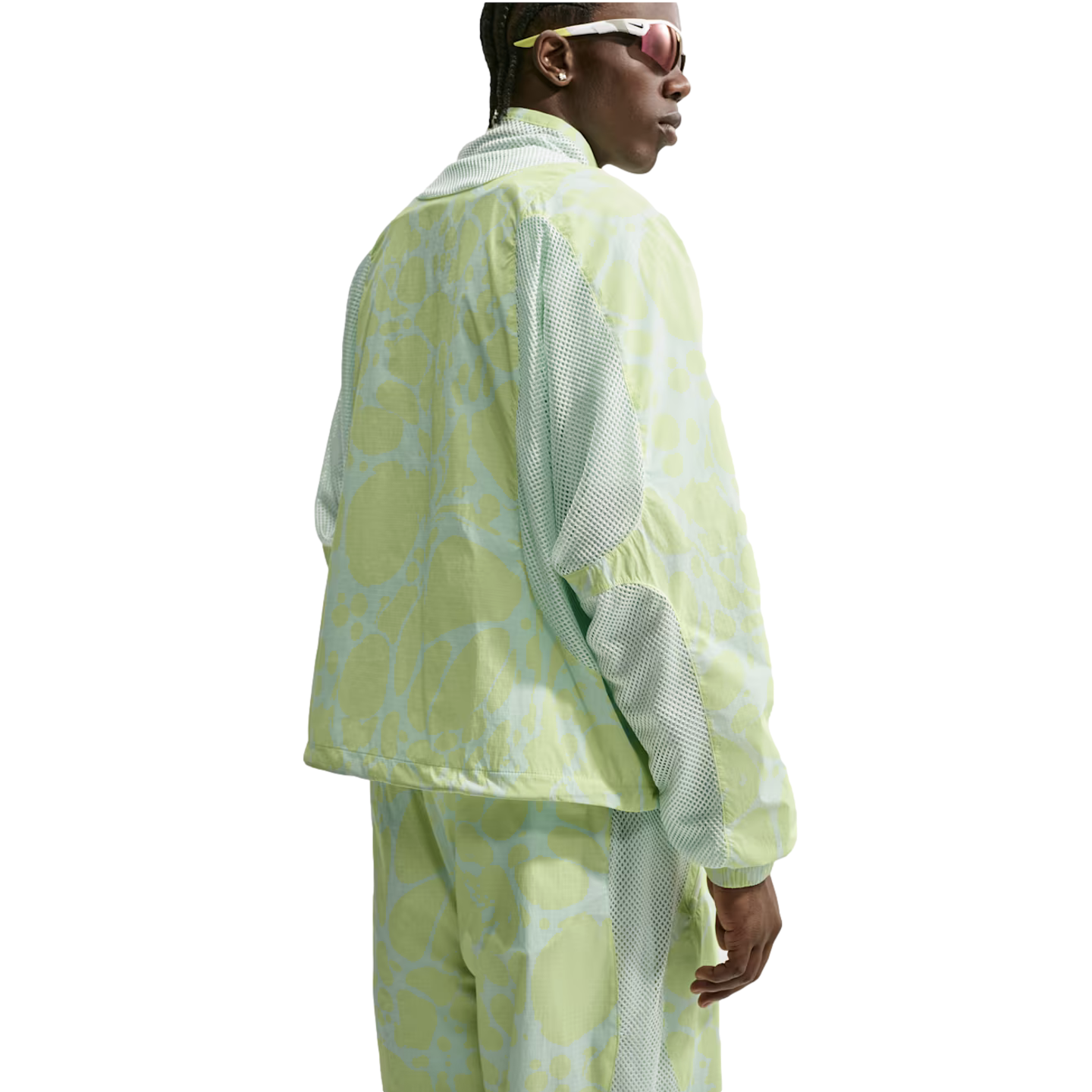 A person wearing a lime green and light blue abstract patterned tracksuit with mesh panels and sporty sunglasses, seen from the back against a white background.