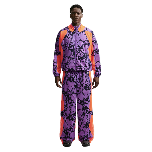 A Black man models a vibrant purple tracksuit with black marble patterns and bright orange mesh side panels, standing against a white background.