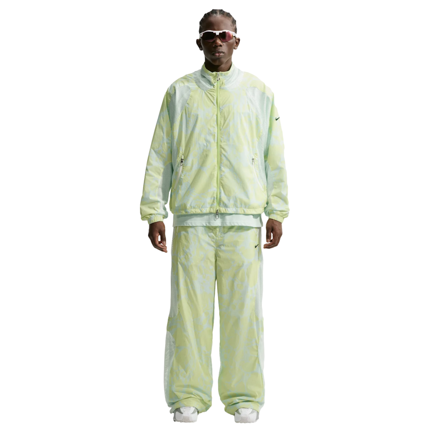 A person wearing a light green and white patterned Nike tracksuit, futuristic white sunglasses, and white sneakers against a white background.