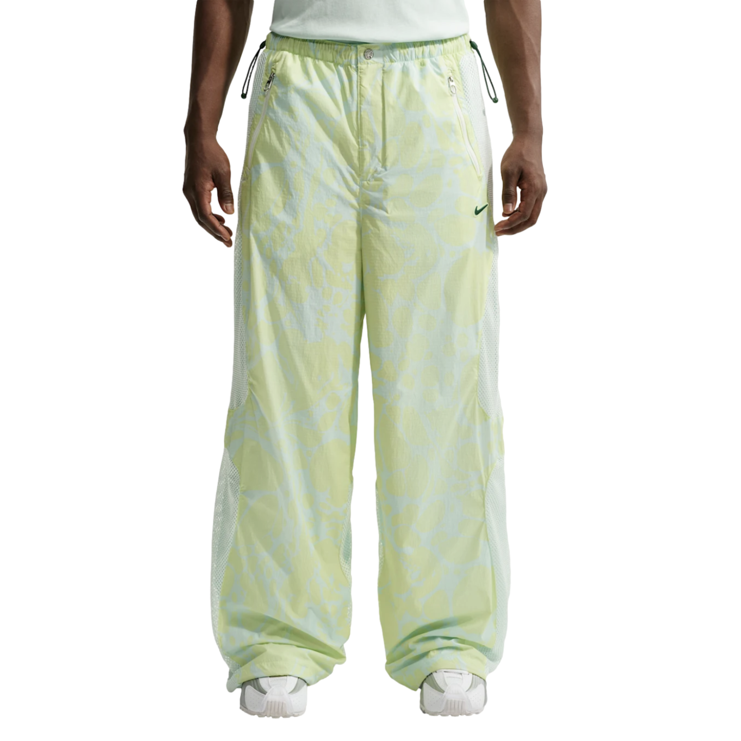 A front view from the waist down of a person wearing baggy, light green and white patterned Nike track pants with white mesh side panels and white sneakers.