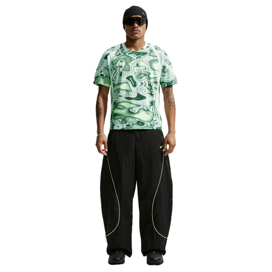 Man wearing a green patterned Nike shirt, baggy black pants with lime yellow piping, a black beanie, and sunglasses against a plain white background.