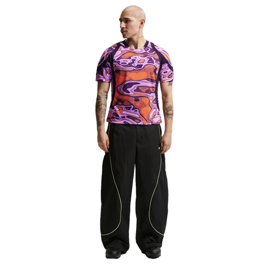A man wearing a purple and orange psychedelic-patterned t-shirt and black wide-leg pants with neon piping, standing against a white background.
