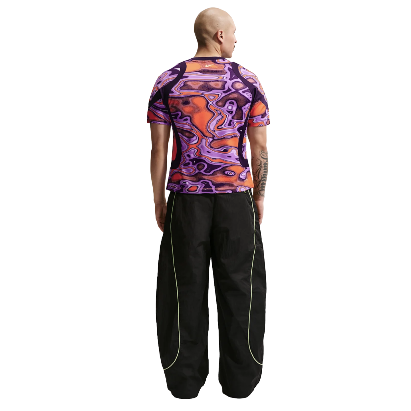 Rear view of a man in a vibrant purple and orange patterned shirt and oversized black track pants with neon green piping, against a plain white background.