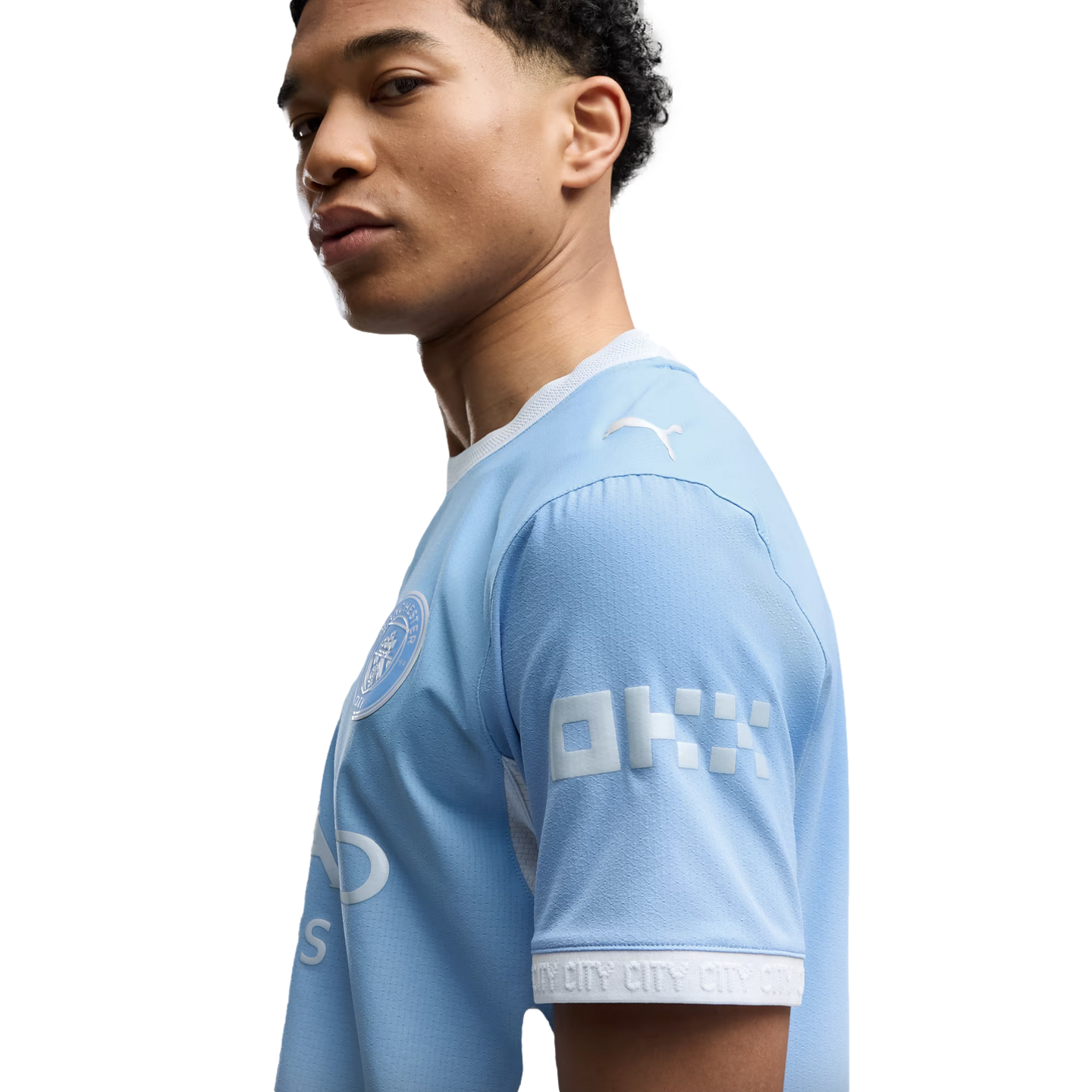 Side profile of a young man with curly hair wearing a light blue Manchester City football jersey against a white background.