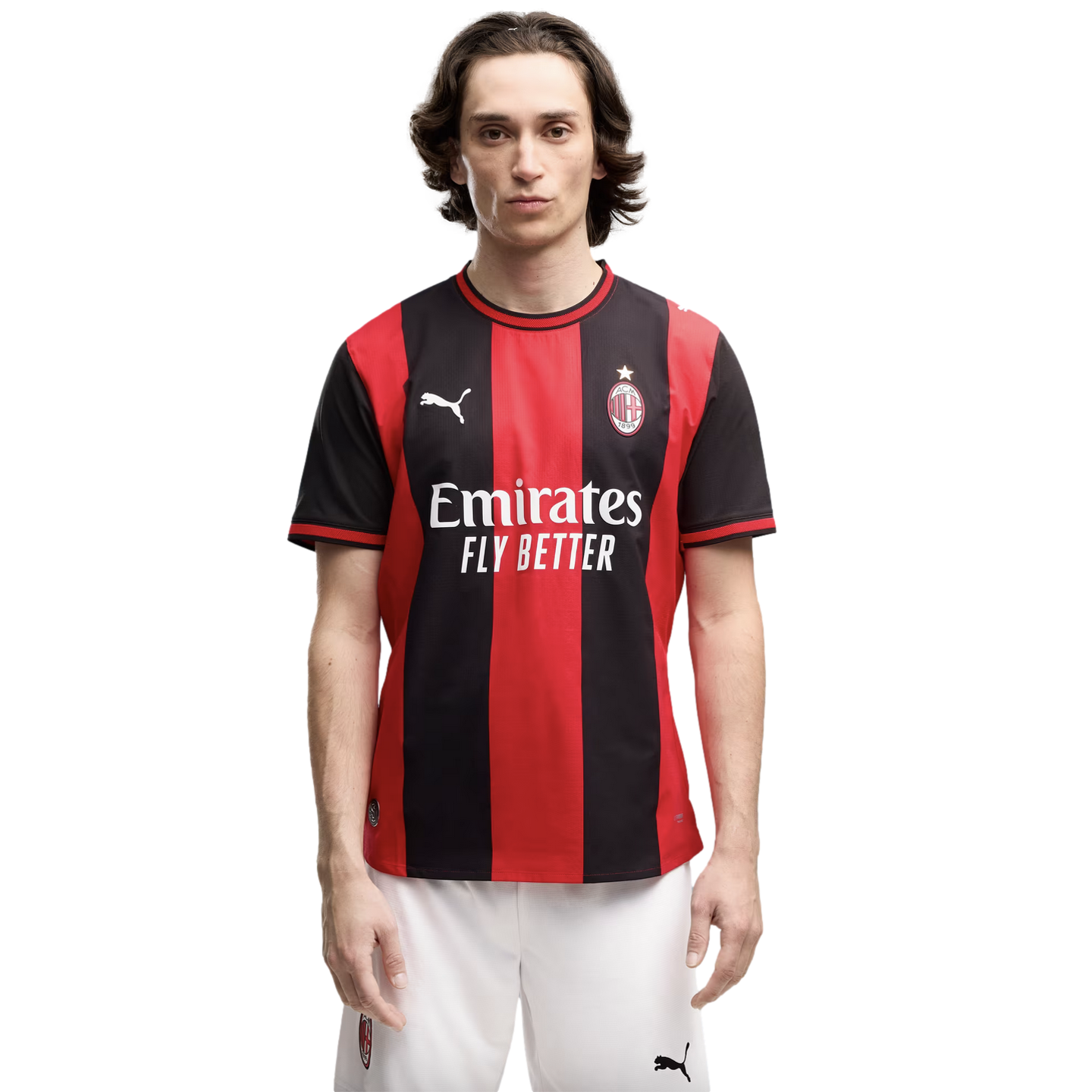 A man with long dark hair wearing a red and black striped AC Milan soccer jersey and white shorts, standing against a plain white background.