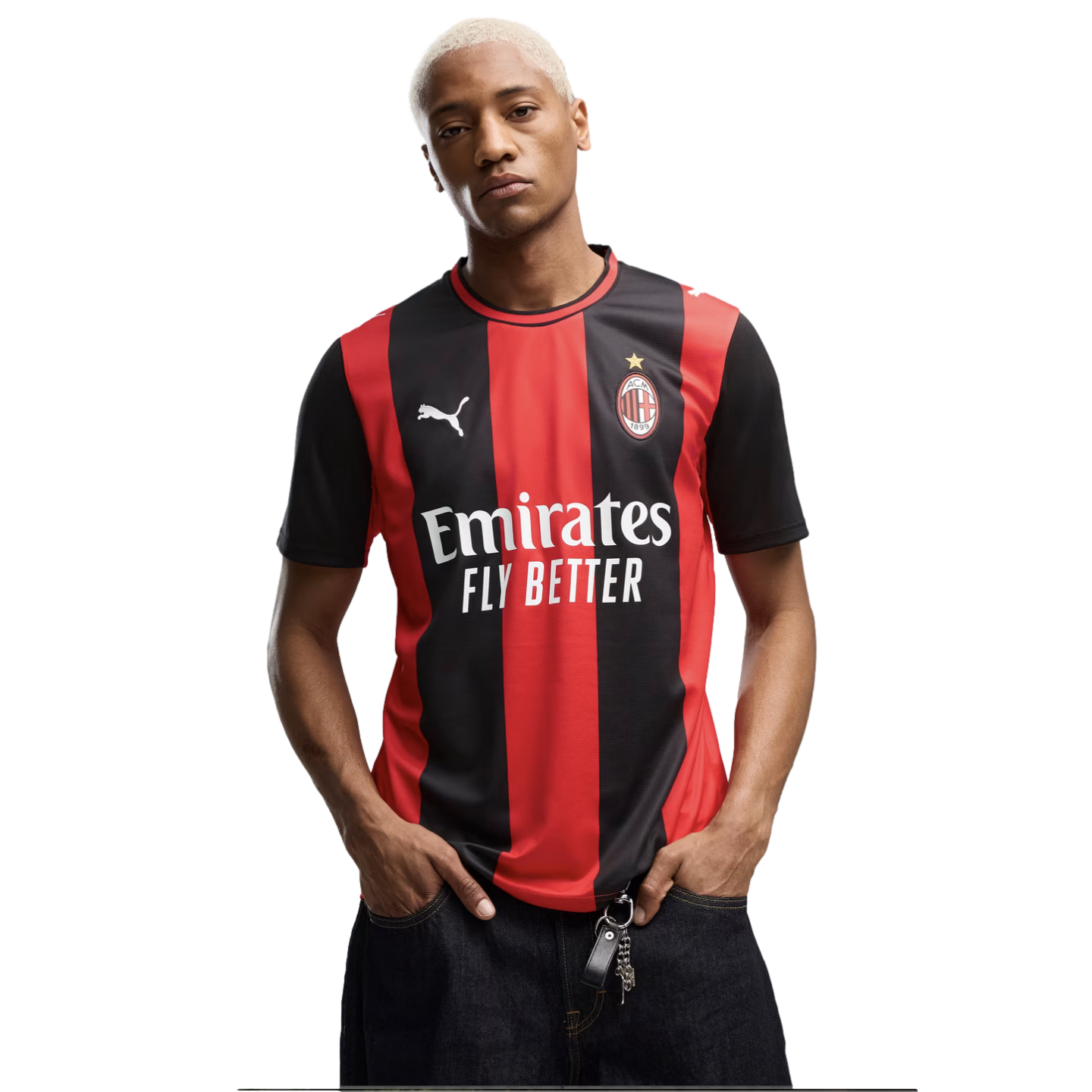 A man with short blonde hair wears a red and black AC Milan jersey and dark jeans, posing with hands in his pockets against a white background.